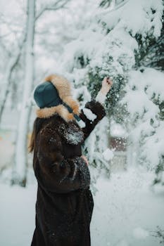 Woman in fur coat and hat plays with snow on an evergreen tree in a winter landscape.