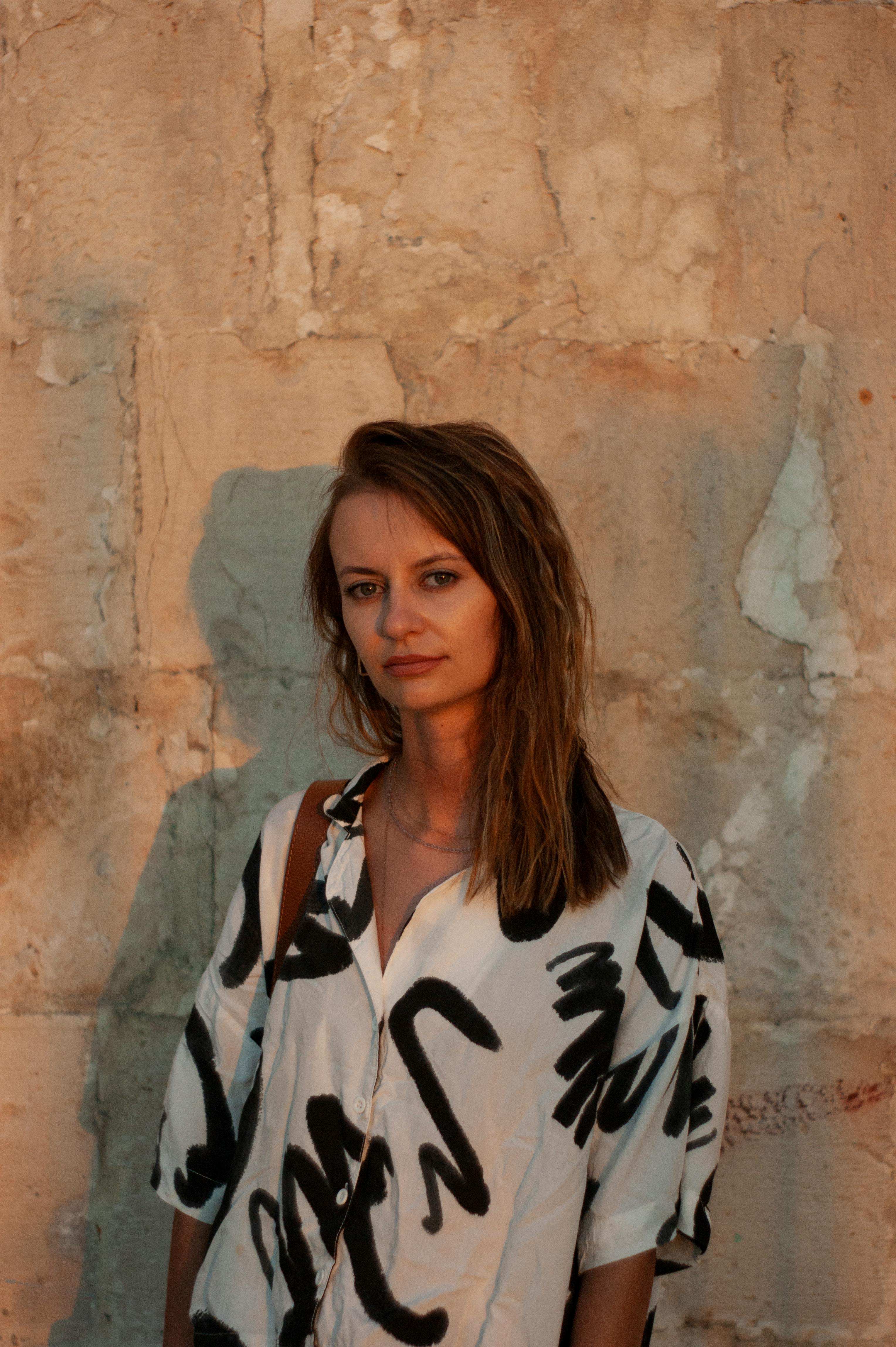 Portrait of a woman standing against a textured stone wall in warm light.