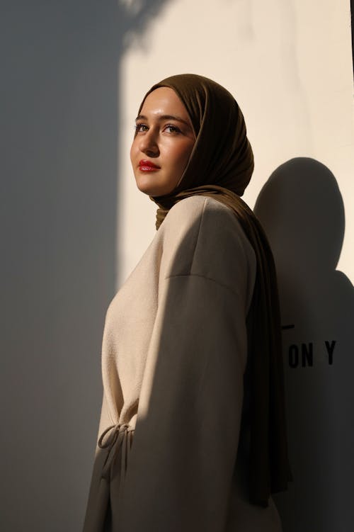 Young Woman in an Elegant Outfit and Hijab Posing in Sunlight against a White Wall