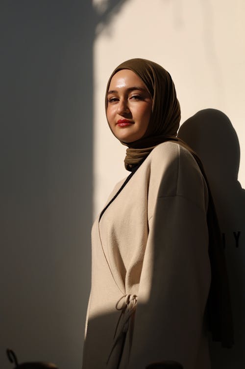Young Woman in an Elegant Outfit and Hijab Posing in Sunlight against a White Wall
