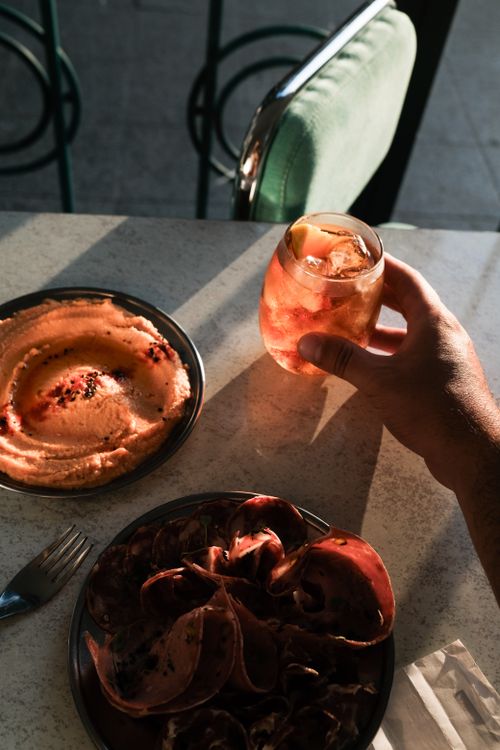Free A person enjoys a cocktail with hummus and cold cuts on a sunny patio table. Stock Photo