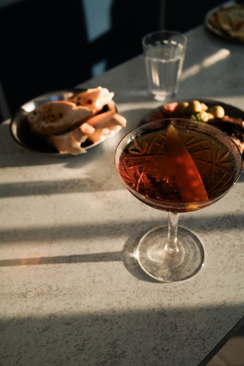 Free A stylish cocktail with an orange twist, bread slices, and olives on a sunlit table. Stock Photo