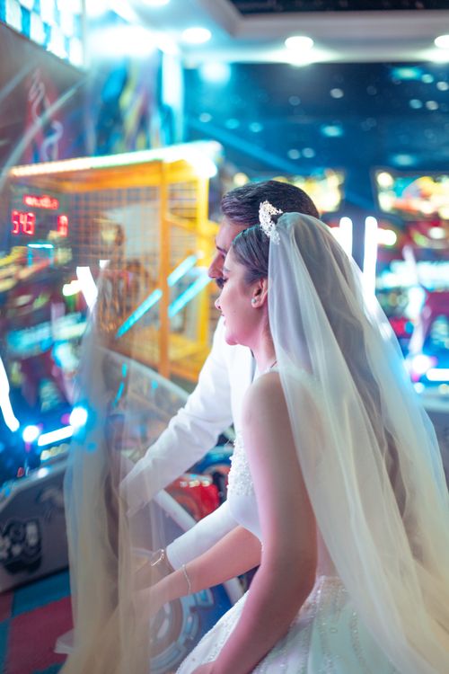 Free A couple in wedding attire enjoys playing arcade games, capturing a unique and playful moment. Stock Photo