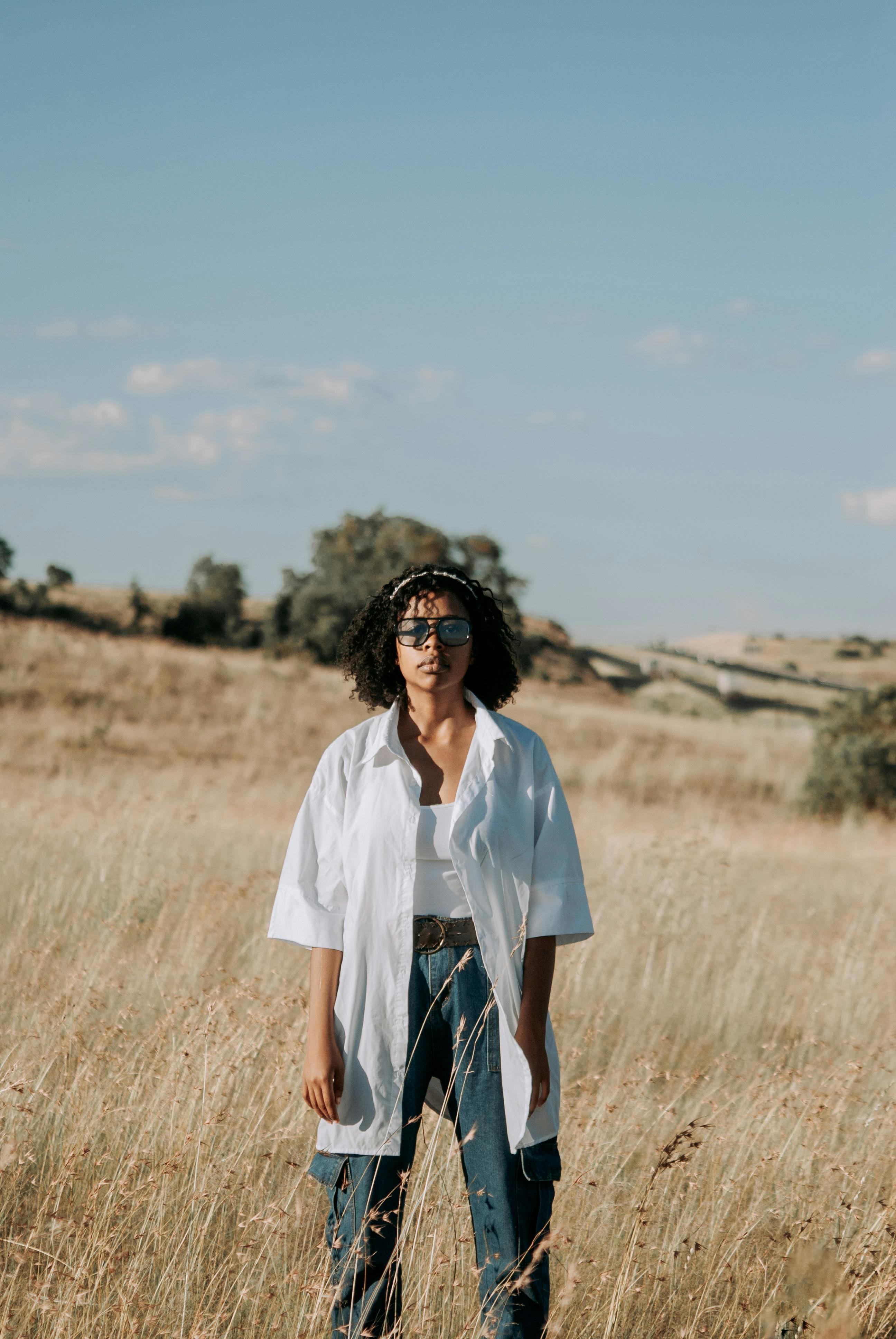Fashionable woman stands confidently in a sunny meadow wearing a white shirt and jeans.