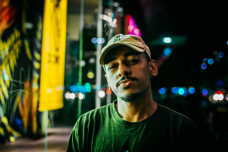 Man with facial hair wearing a cap standing against vibrant city nightlife backdrop.