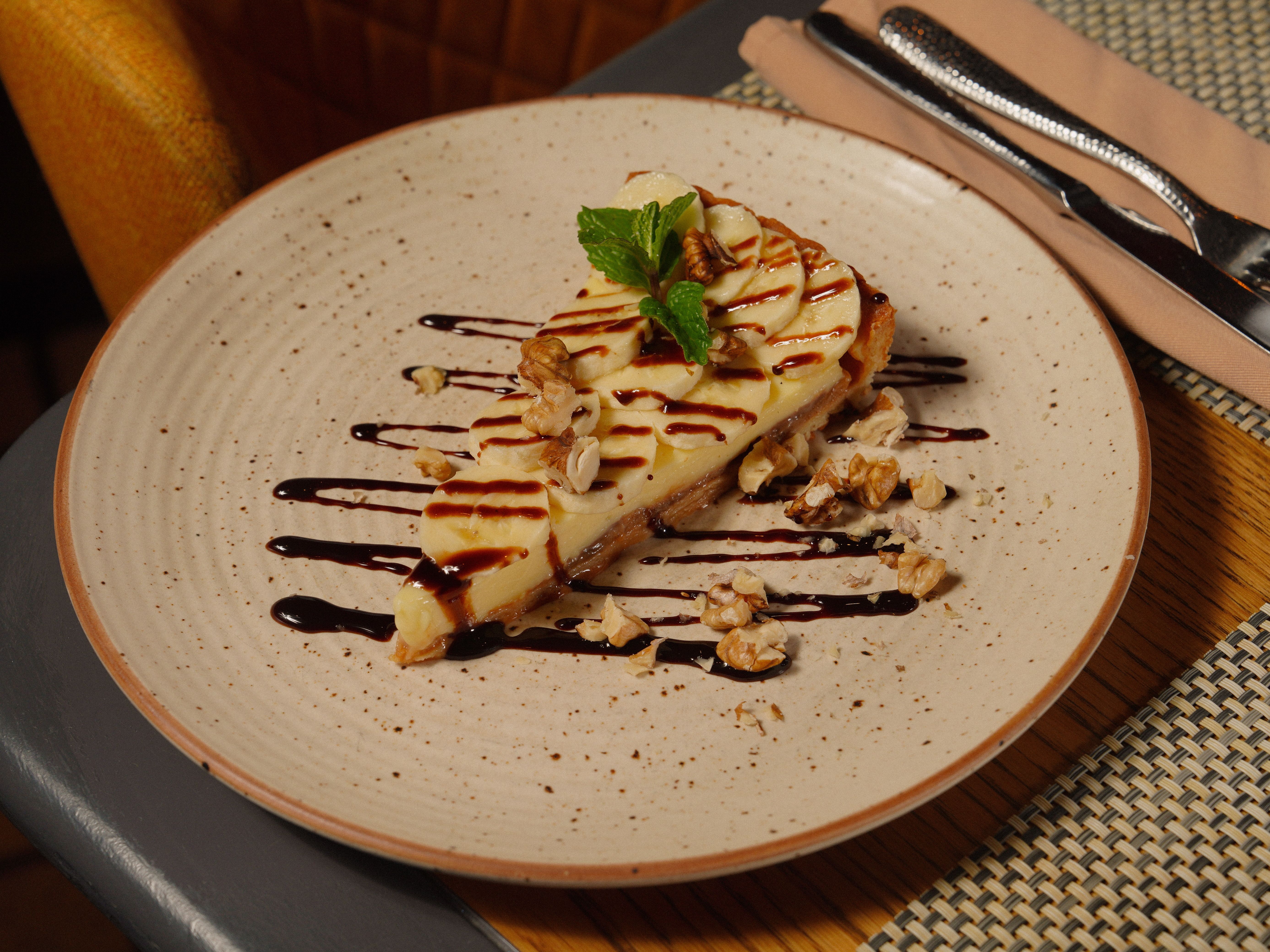 A slice of creamy cheesecake topped with nuts and mint, drizzled with chocolate sauce.