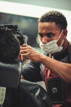 Barber in face mask trims client's hair in a modern shop, focusing intently.