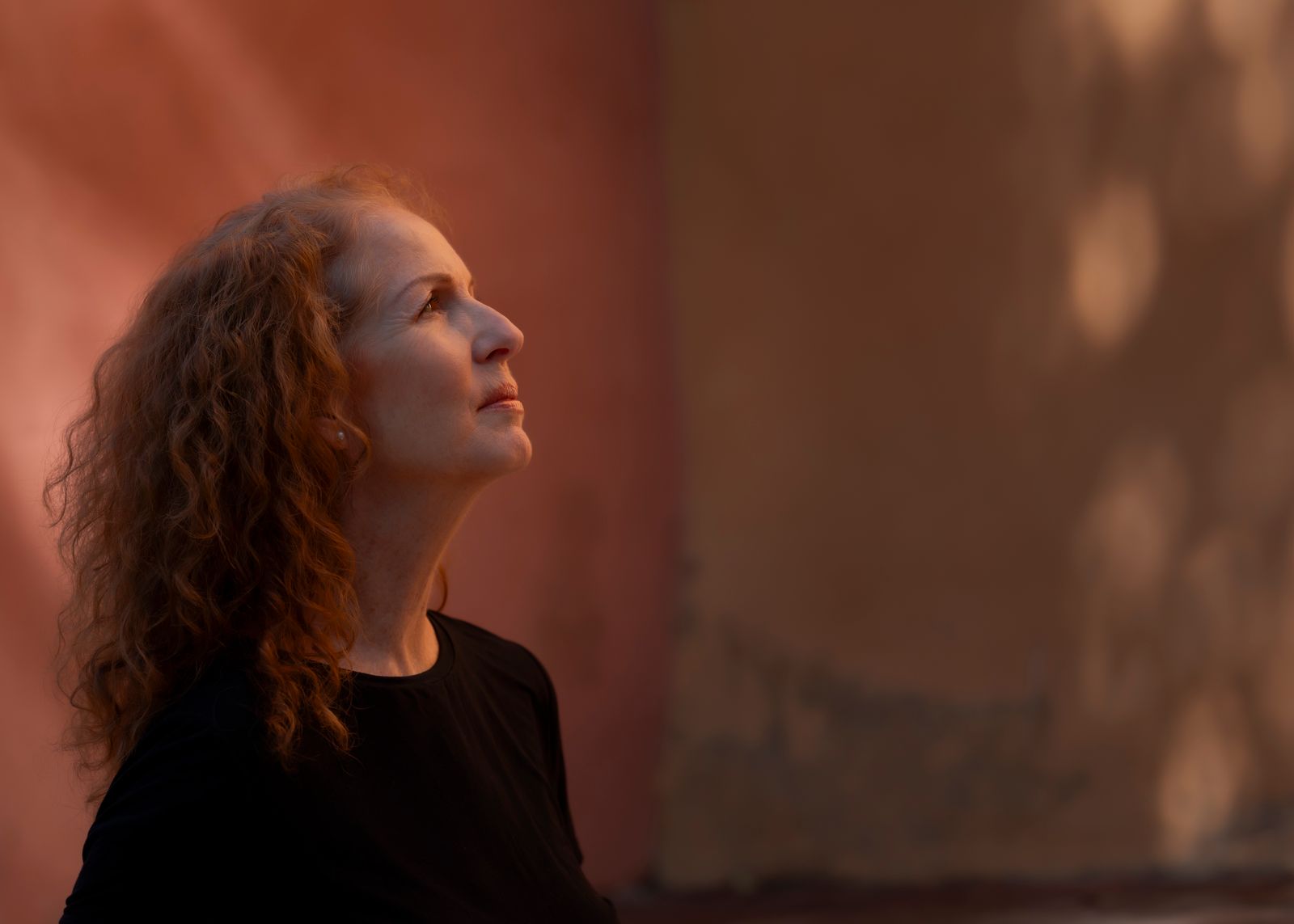 Woman with curly red hair in a black top looks upward in warm light against a terracotta wall