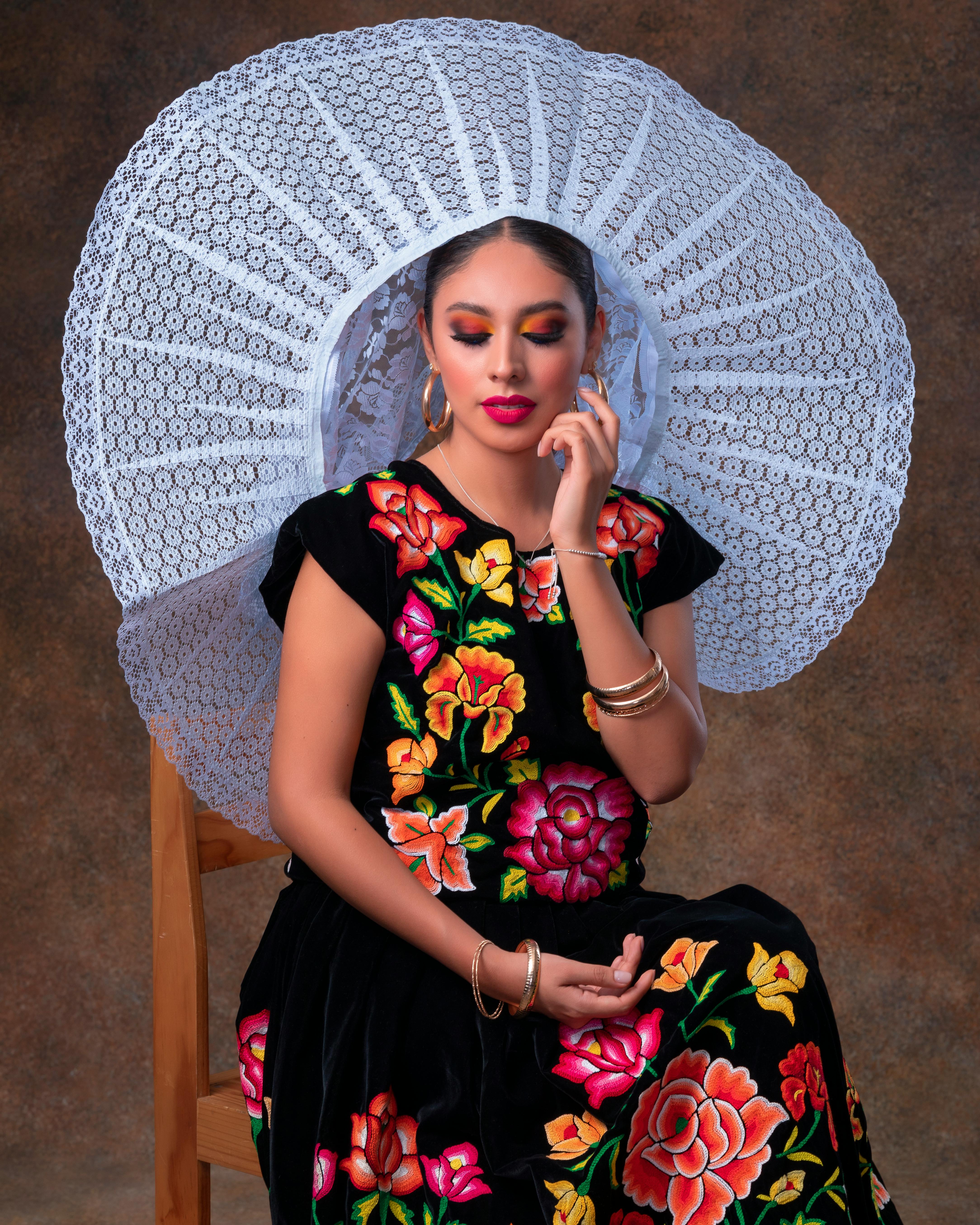 Elegant portrait of a woman in vibrant traditional dress with floral embroidery, wearing a large lace hat, seated indoors.