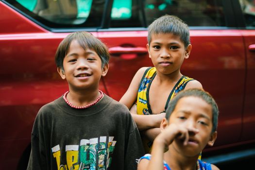 Three children standing together and smiling in front of a red car outdoors.