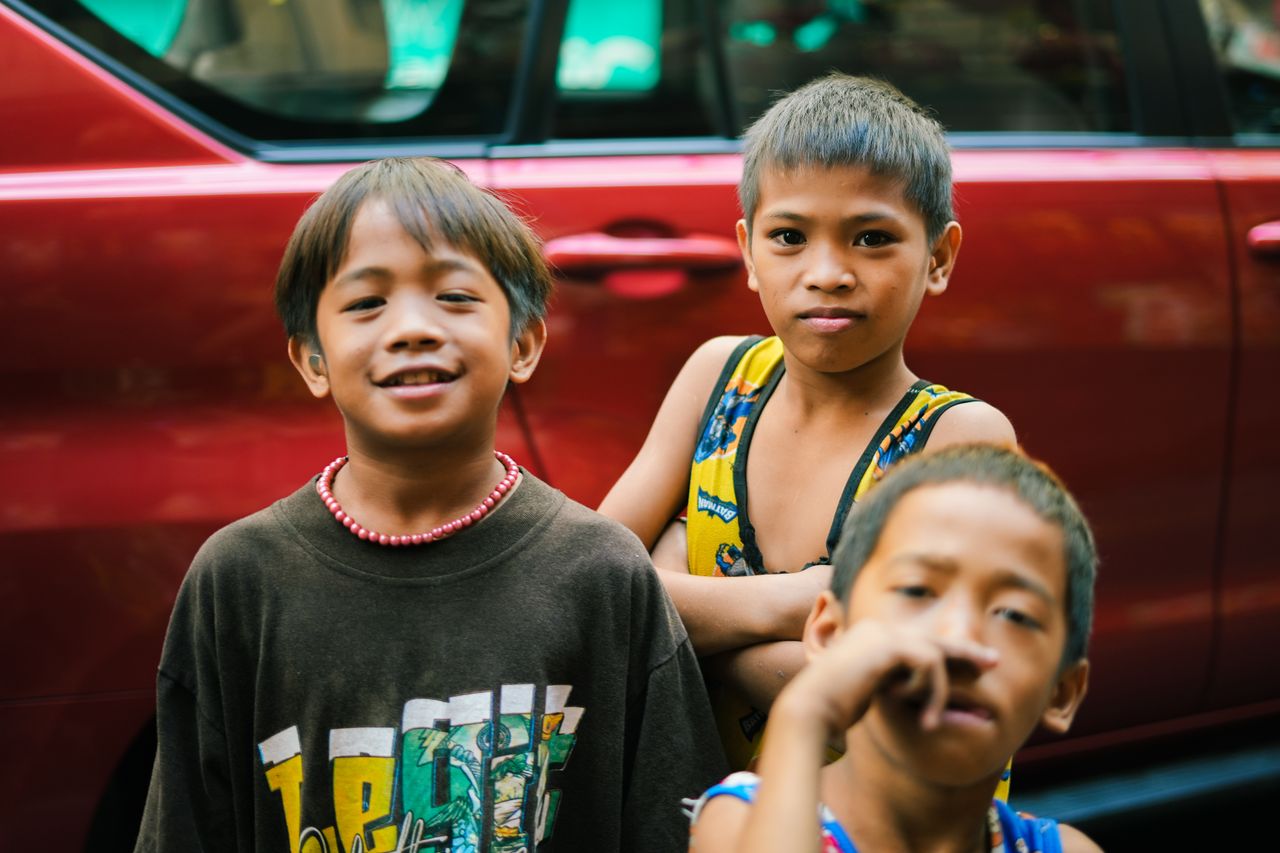 three children standing together and smiling in front of a red car out