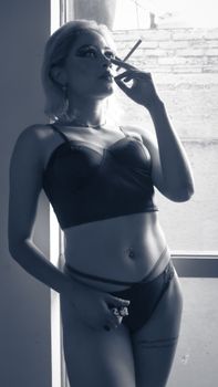 Artistic portrait of a woman in lingerie, smoking by a window. Black and white photography style.