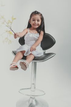 Young girl sitting on a modern chair with a leaf branch, smiling in a studio setting.