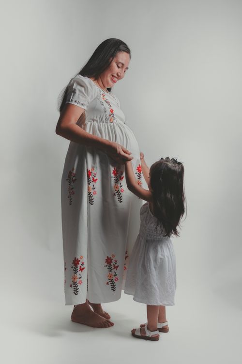 Free A pregnant mother in a floral dress stands gracefully with her young daughter in a studio setting. Stock Photo