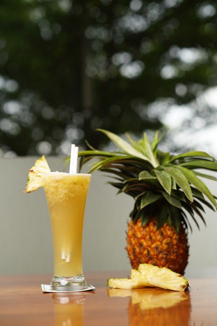 A tall glass of golden pineapple juice with ice cubes, set beside a halved fresh pineapple and its green crown on a wooden board.