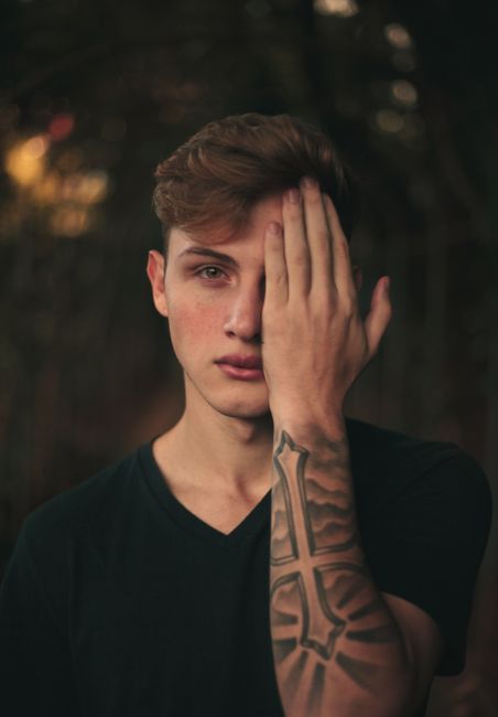 A tattooed man in a black t-shirt poses with one hand covering his eye in a moody, artistic portrait.