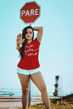 在一个阳光明媚的日子里，一位时尚的女人穿着红色衬衫，以自信的姿势站在停止标志旁，显得格外引人注目