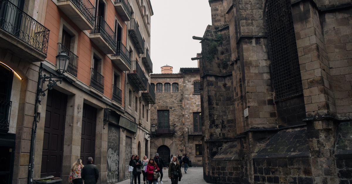 A narrow lane in central Barcelona with people walking between old buildings.
