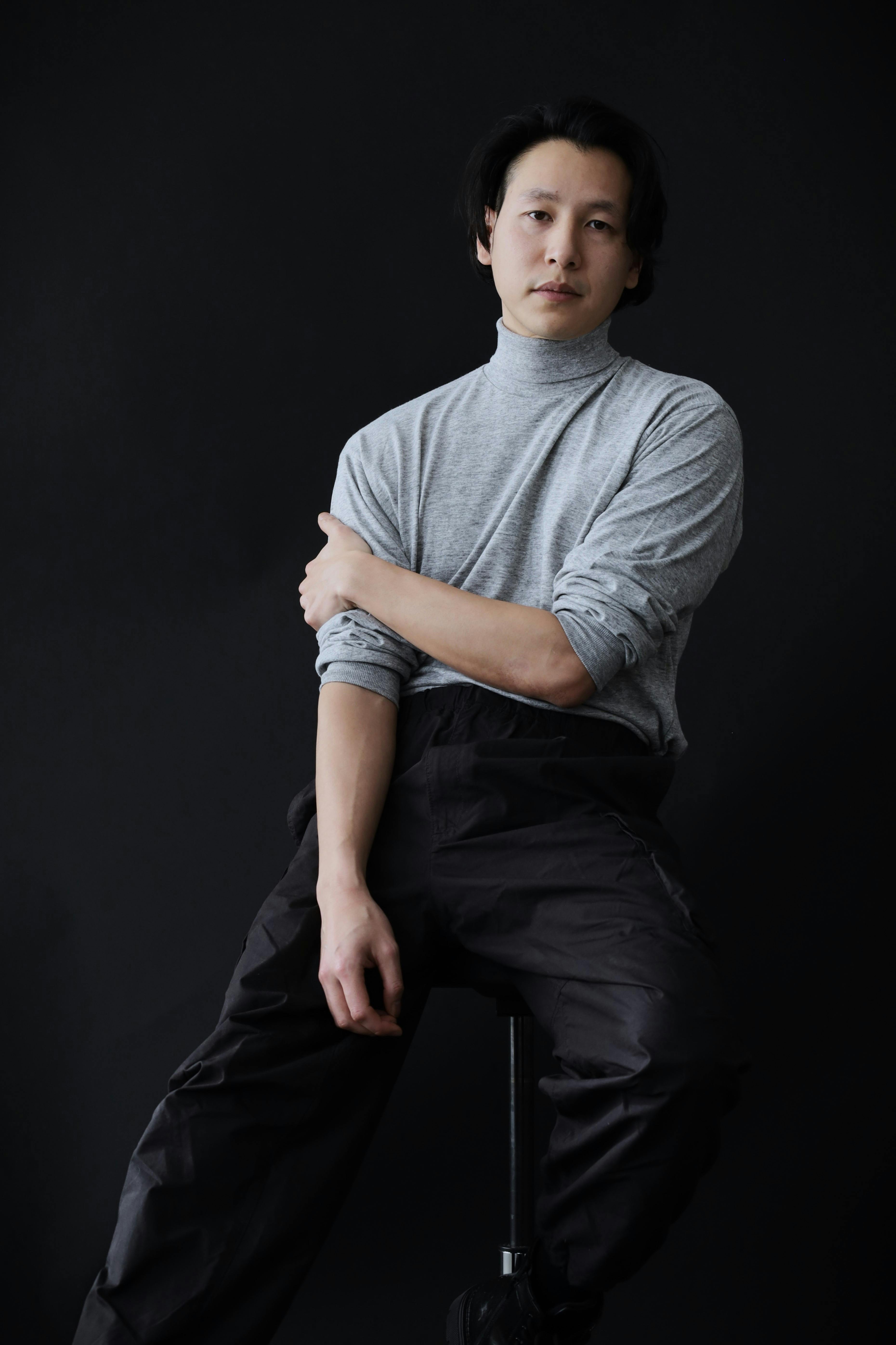 Elegant studio portrait of a seated Asian man in a light gray turtleneck against a black background.