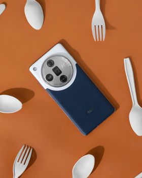 A sleek smartphone displayed on an orange background with scattered white cutlery, captured in a top view.