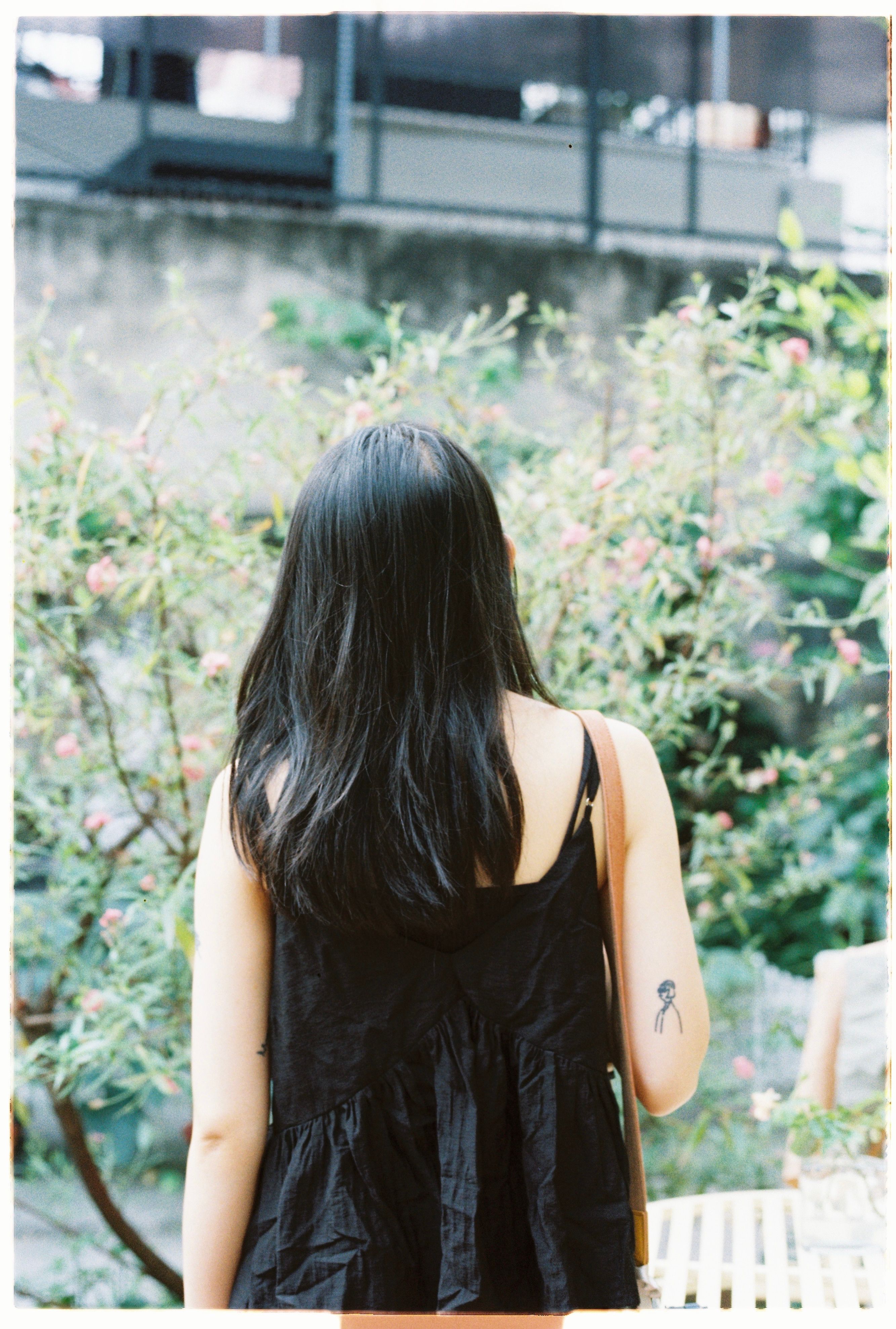 A brunette woman with a tattoo stands in a vibrant garden surrounded by greenery and pink flowers.