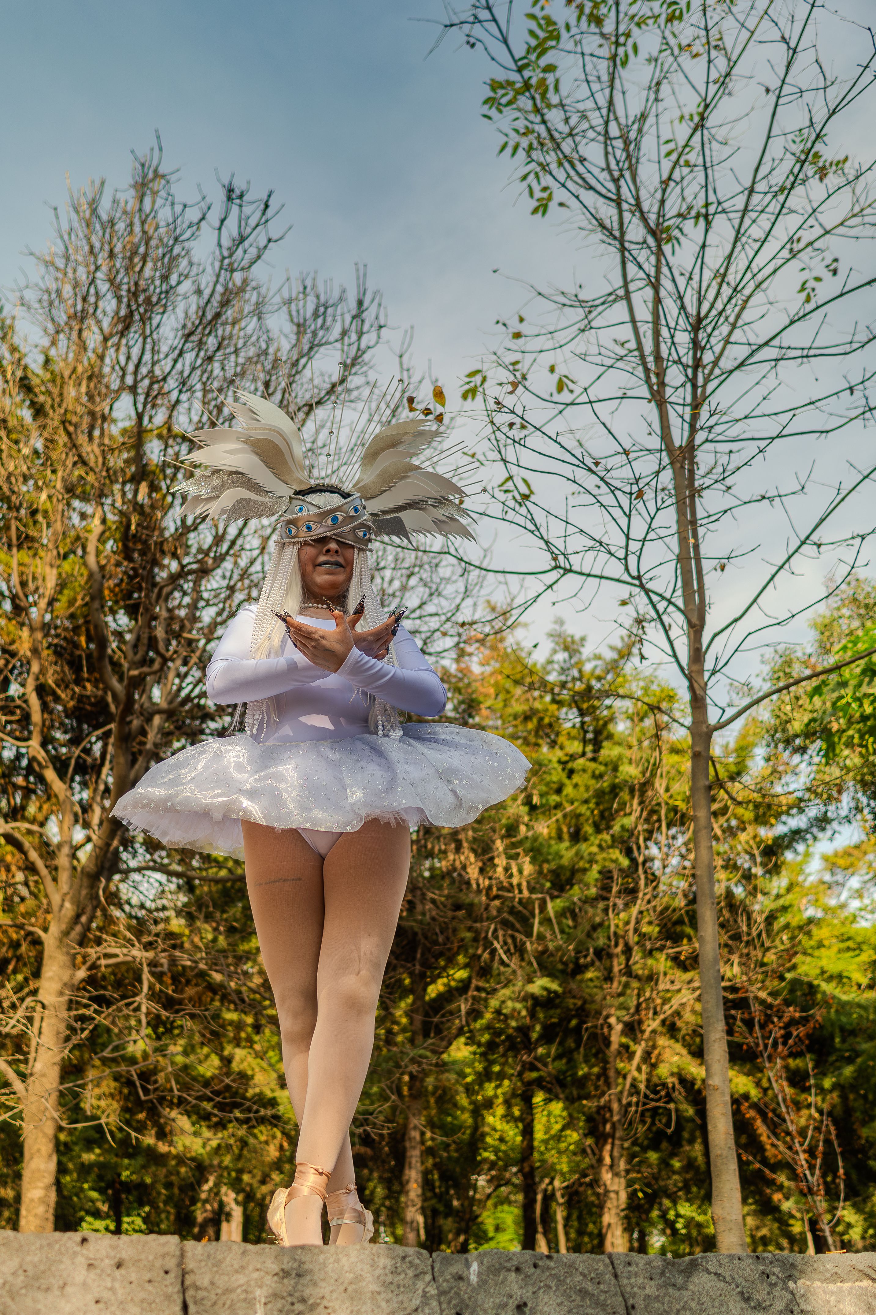 Elegant woman in angelic attire poses outdoors amid lush trees.