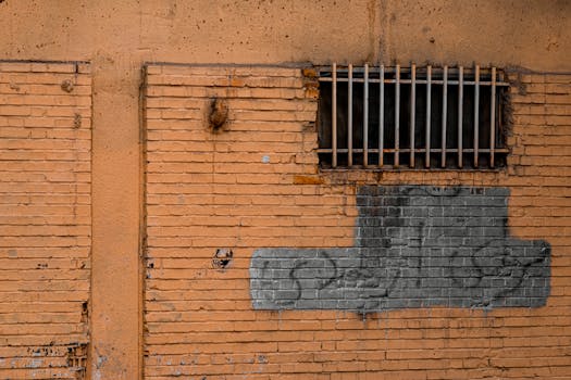 An abandoned brick wall with a barred window and graffiti in an urban area.