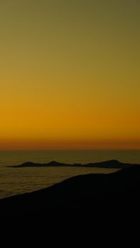 A scenic coastal sunset featuring silhouettes of hills against a vibrant yellow sky.