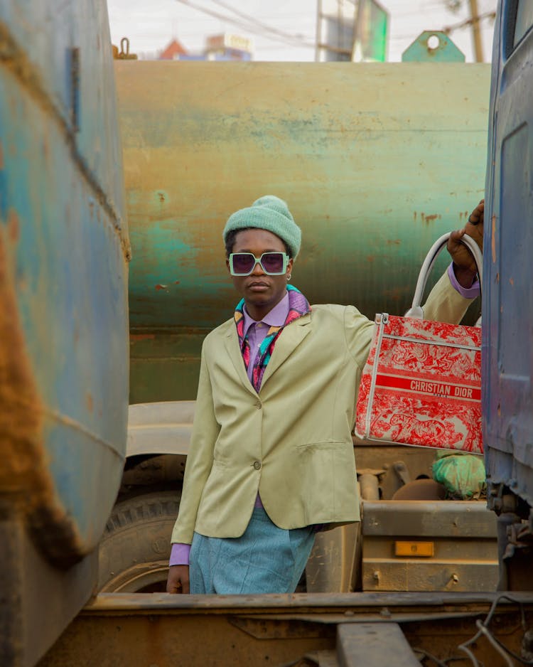 Woman In A Colorful Outfit And Sunglasses, Holding A Purse And Standing Between Rusty Trucks 