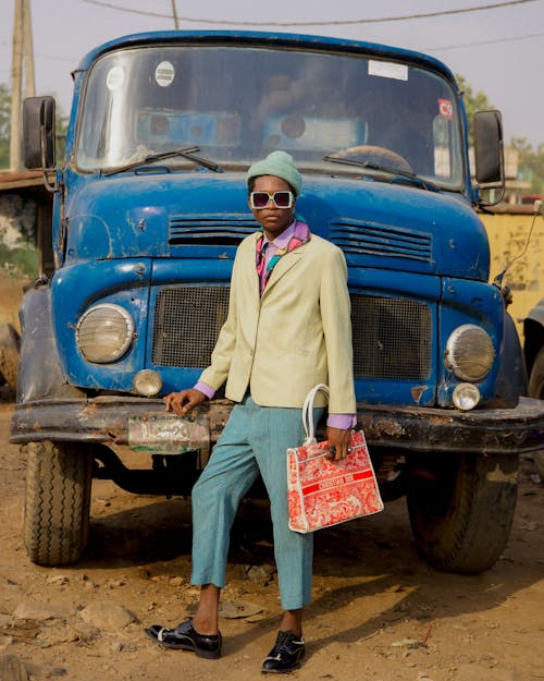 Woman in a Colorful Outfit and Sunglasses, Holding a Purse and Standing in front of a Rusty Truck