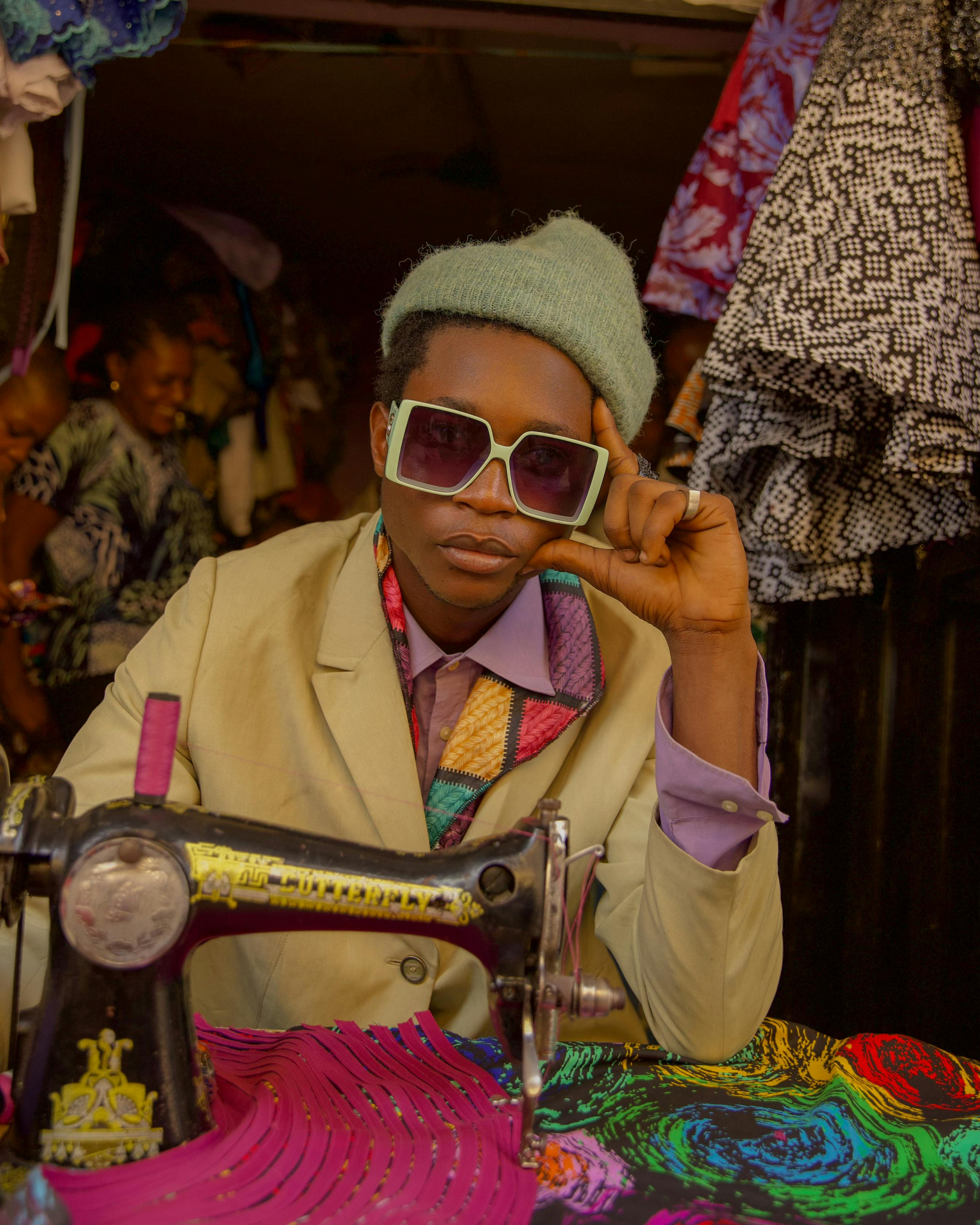 A stylish African woman vendor with sunglasses showcases her sewing craft in a vibrant outdoor market scene.