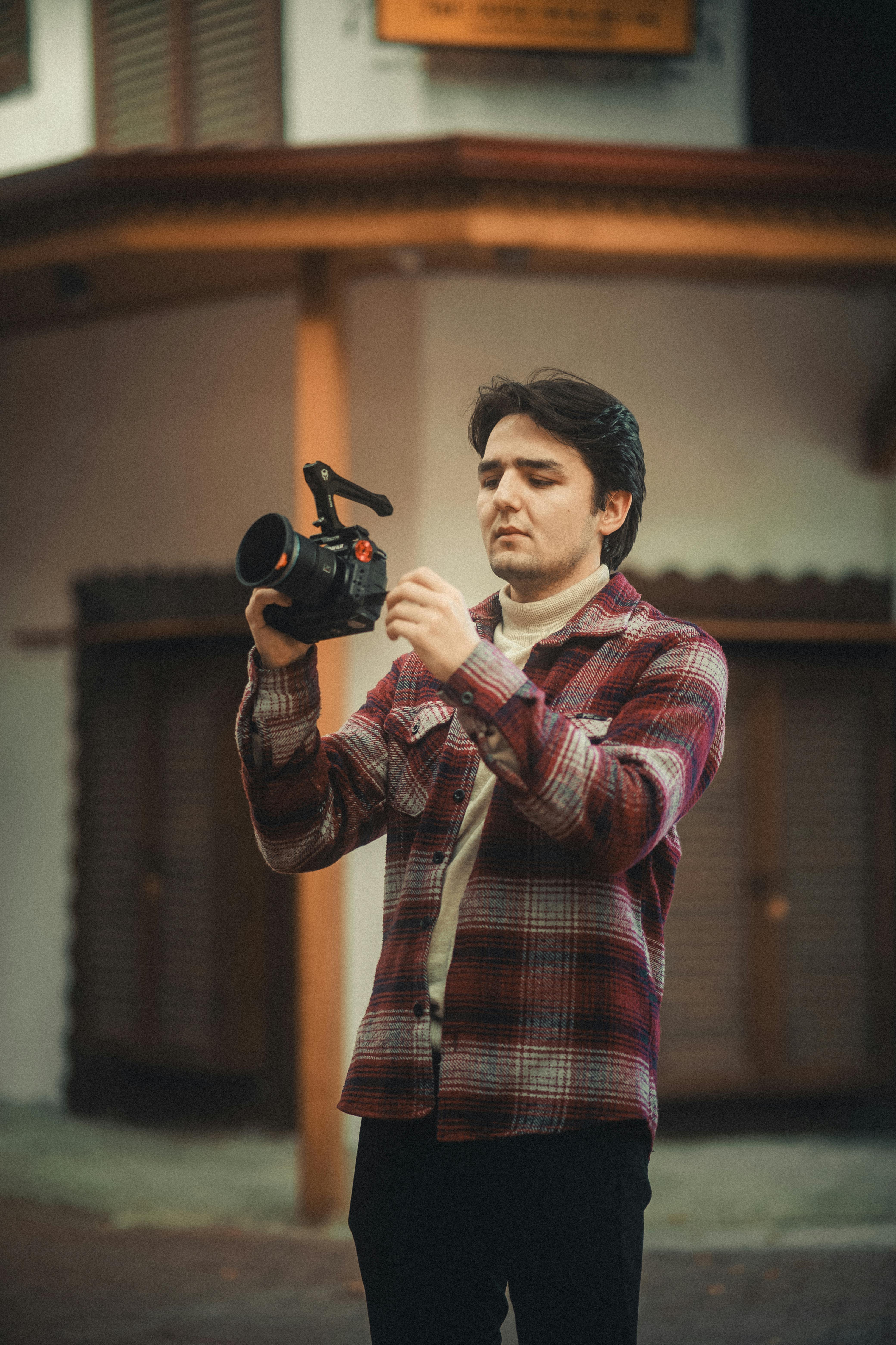 Free A young male photographer adjusting his professional camera outdoors in a casual setting. Stock Photo