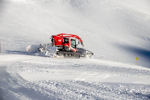 Red snow groomer smoothing snowy slopes in winter mountains during daytime.