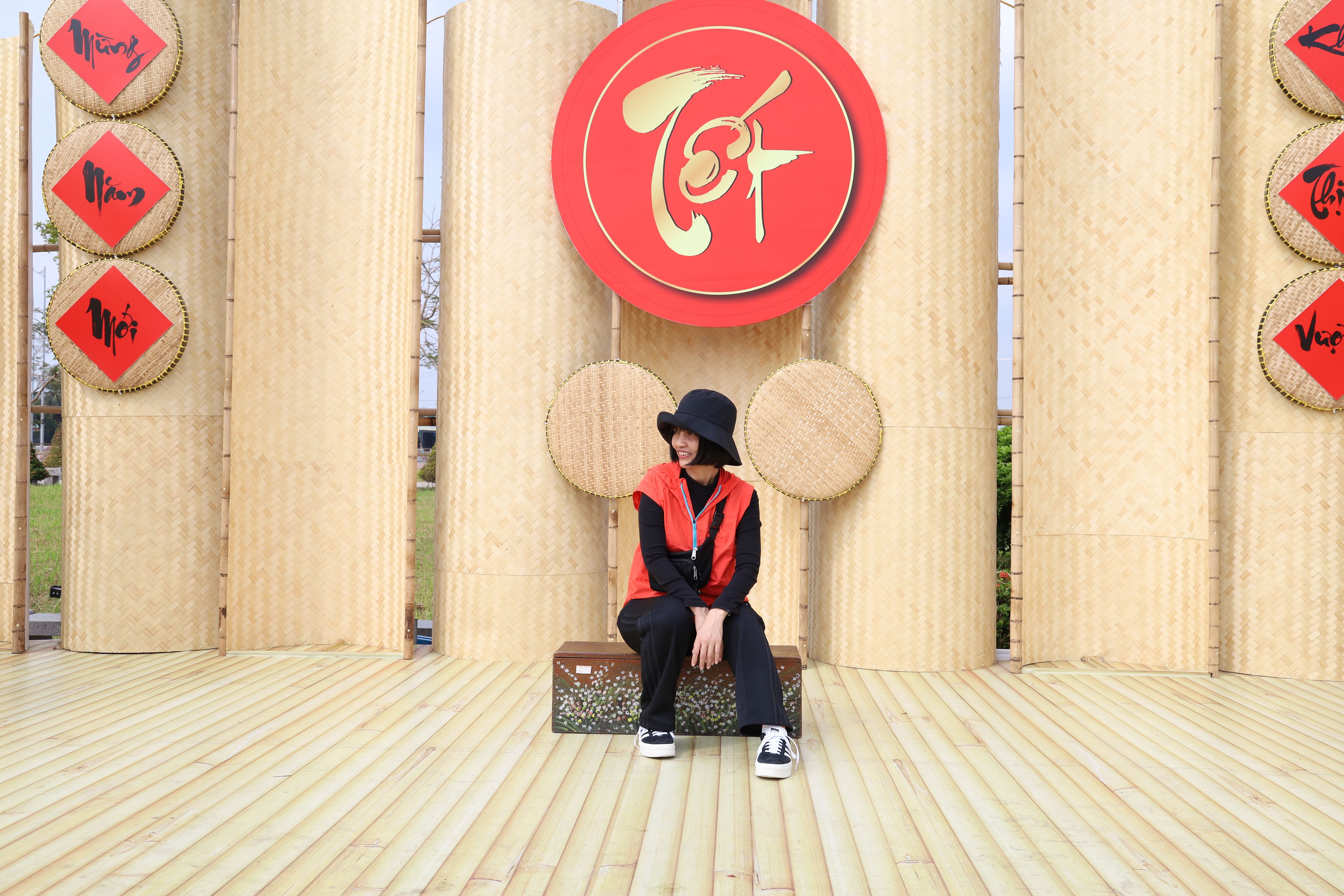 Young Asian woman sitting in front of cultural decorations, wearing a red vest and black hat.