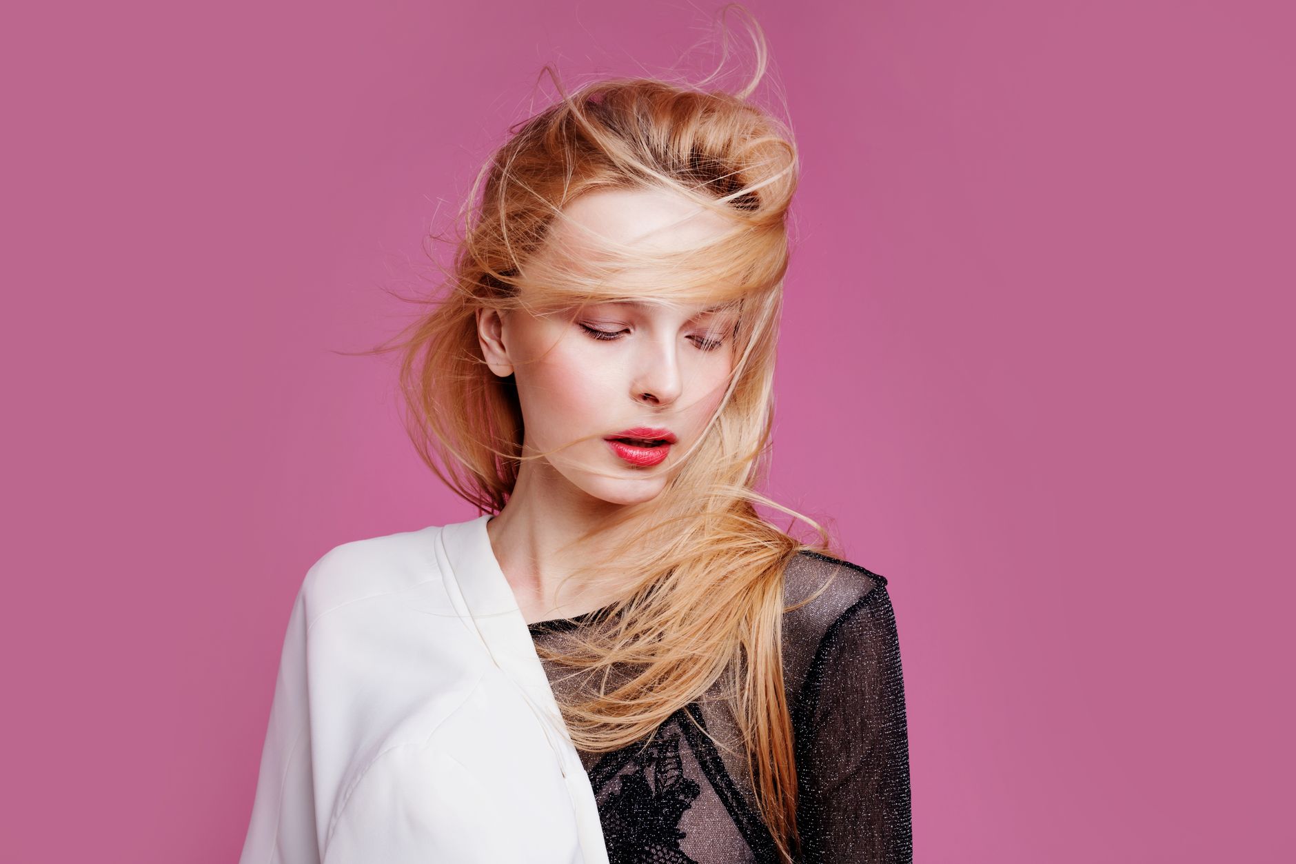 Fashion portrait of a blonde woman with hair blowing gracefully against a pink backdrop, exuding elegance.