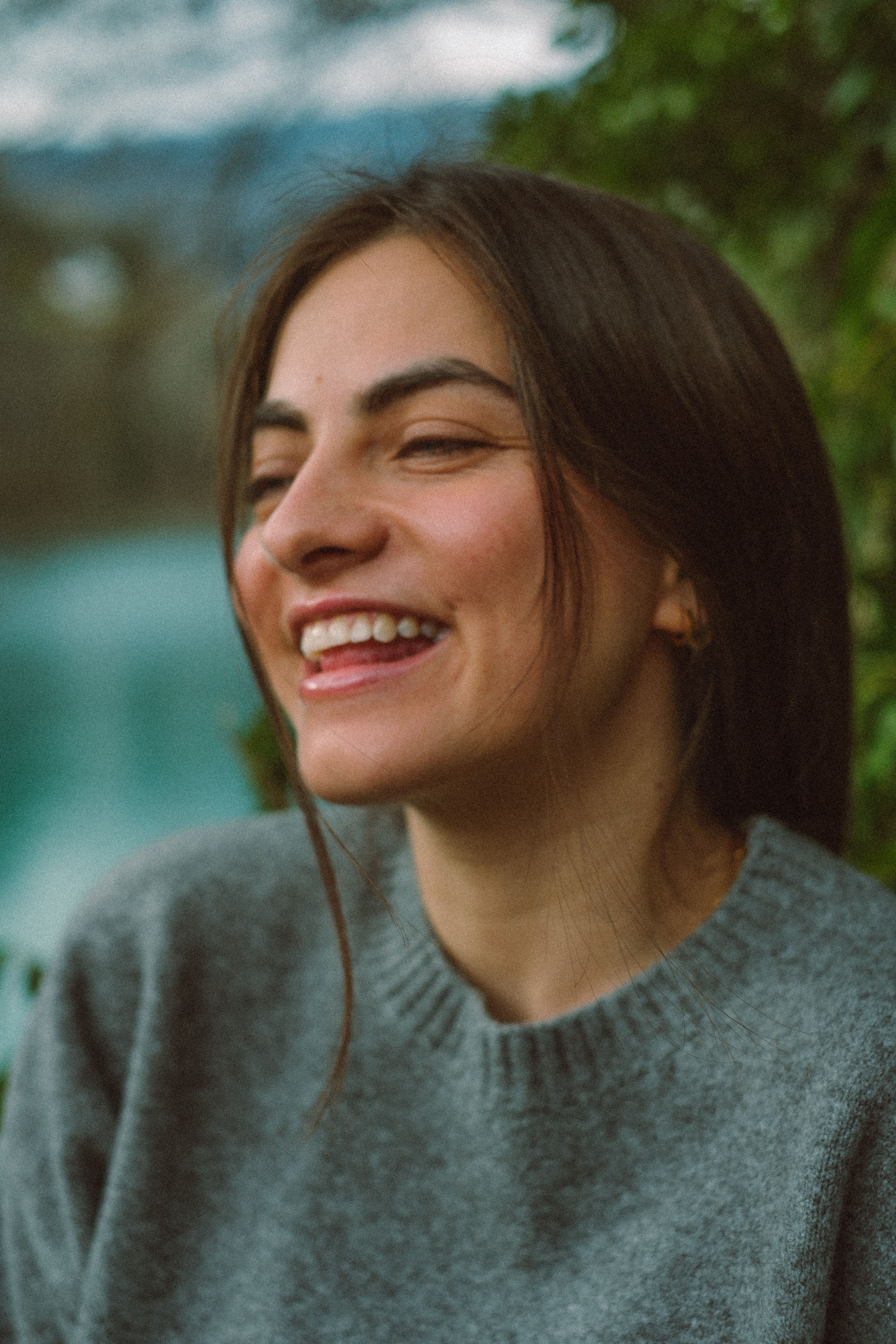 Portrait of a cheerful woman with brown hair, smiling in an outdoor setting, wearing a grey sweater.