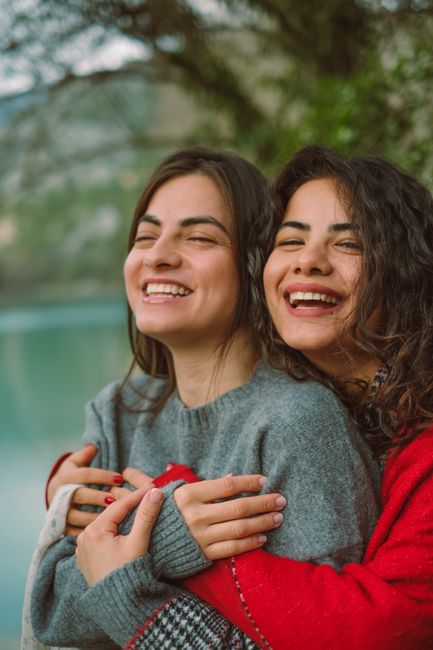 Two women hugging and smiling outdoors beside a serene lake, enjoying a joyful moment.