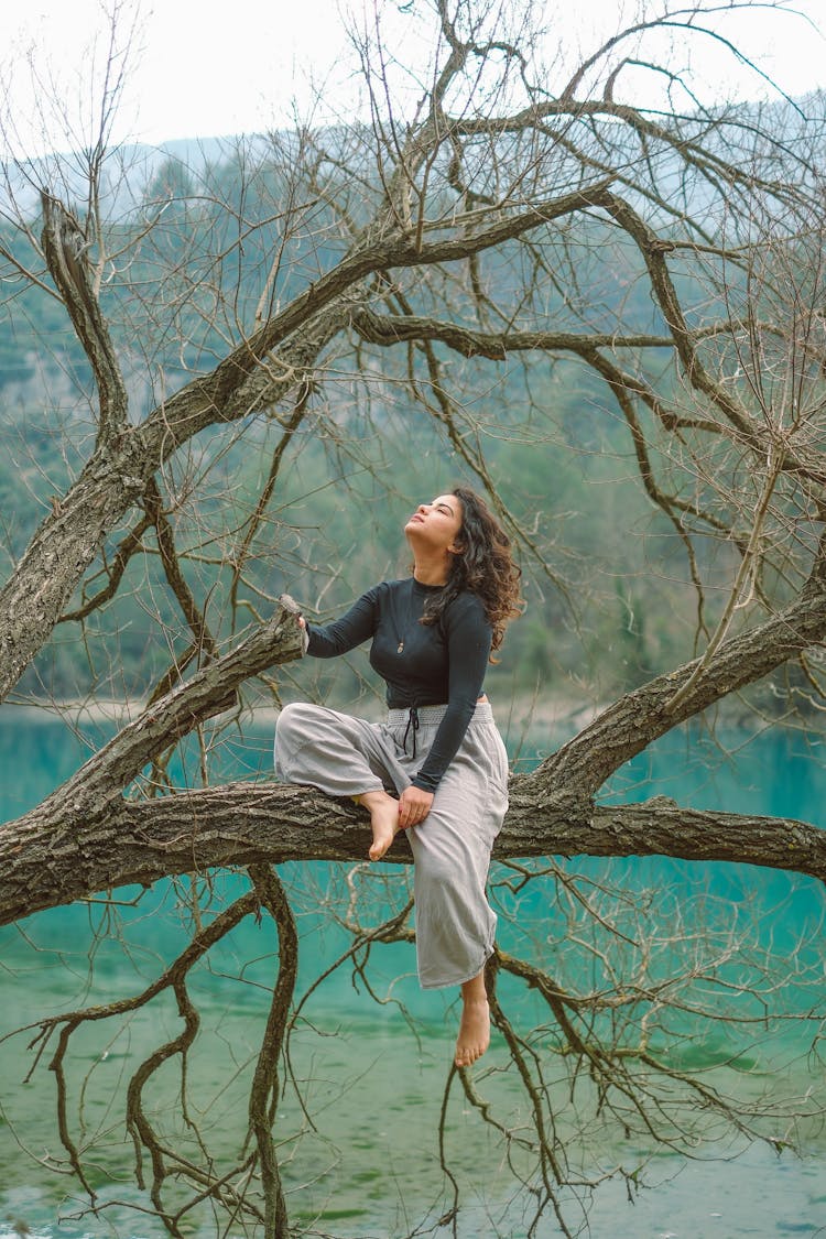 Woman Sitting On A Tree Branch By A Body Of Water 