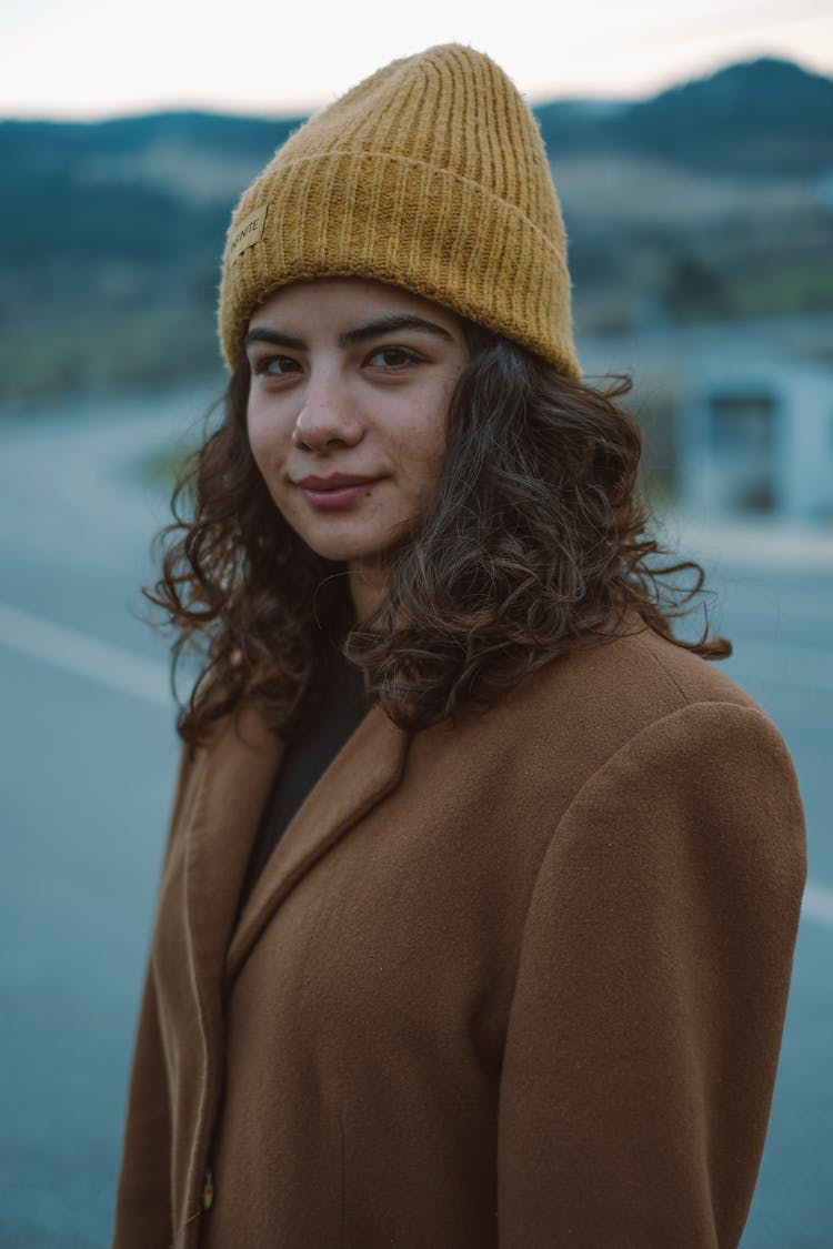 Young Woman In A Brown Coat And Yellow Hat Standing Outside 