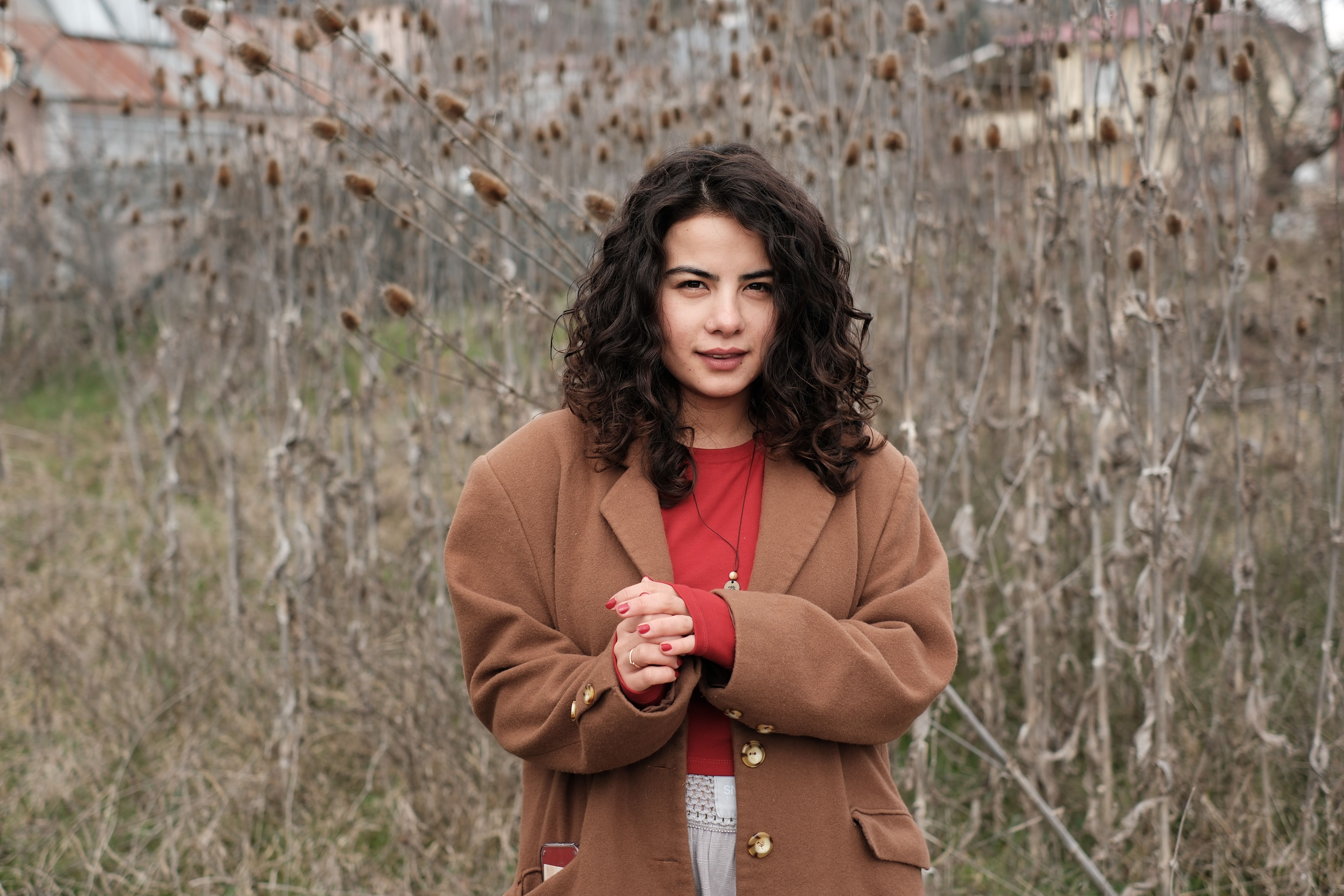 Portrait of a young woman in a brown coat standing outdoors in a rural setting.