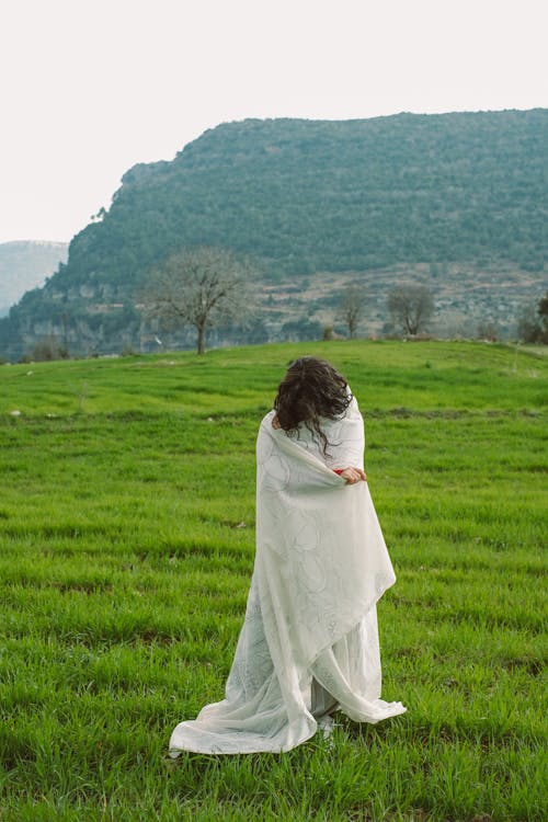 Free Woman Wrapped in a White Blanket Standing on a Meadow Stock Photo