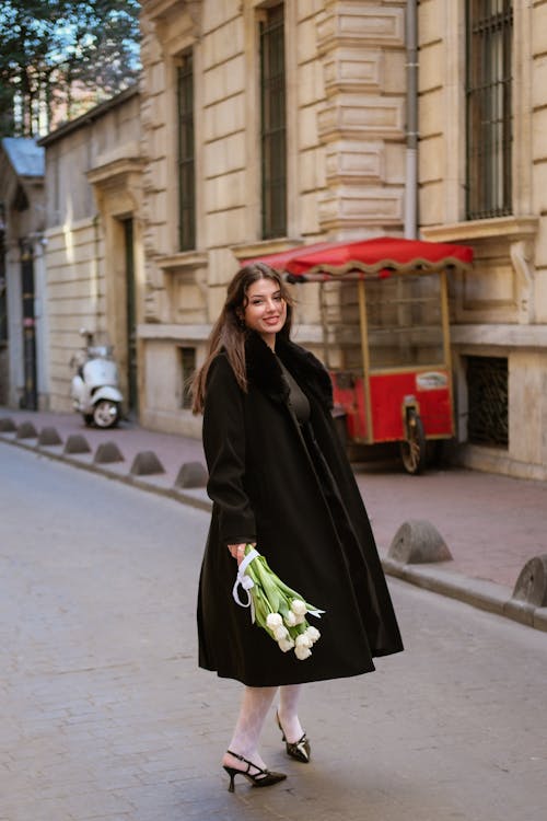 Smiling Woman in Black Coat Standing with Flowers on Street