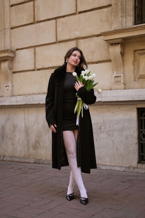 A woman in black coat and white stockings holding a bouquet of flowers