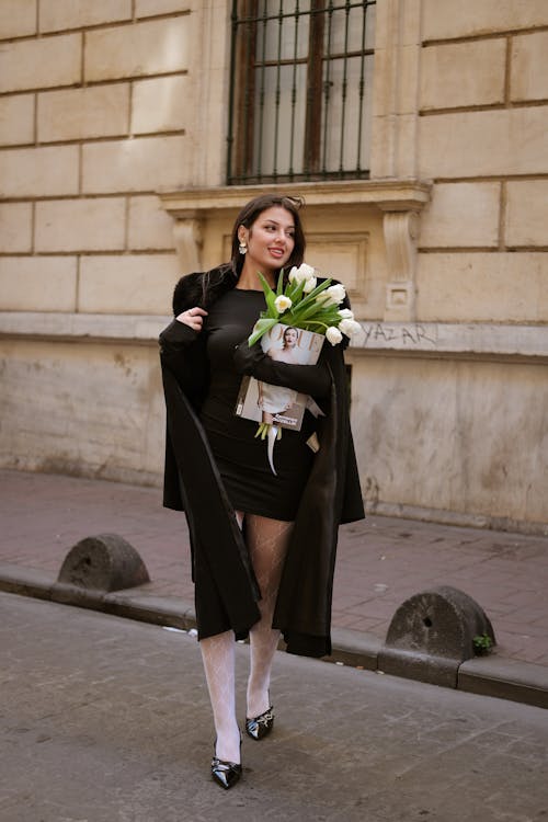 Smiling Woman in Black Coat Walking with Flowers and Magazine on Street