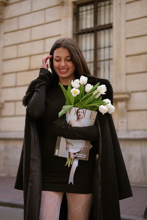 Smiling Woman in Black Coat and with Flowers and Magazine