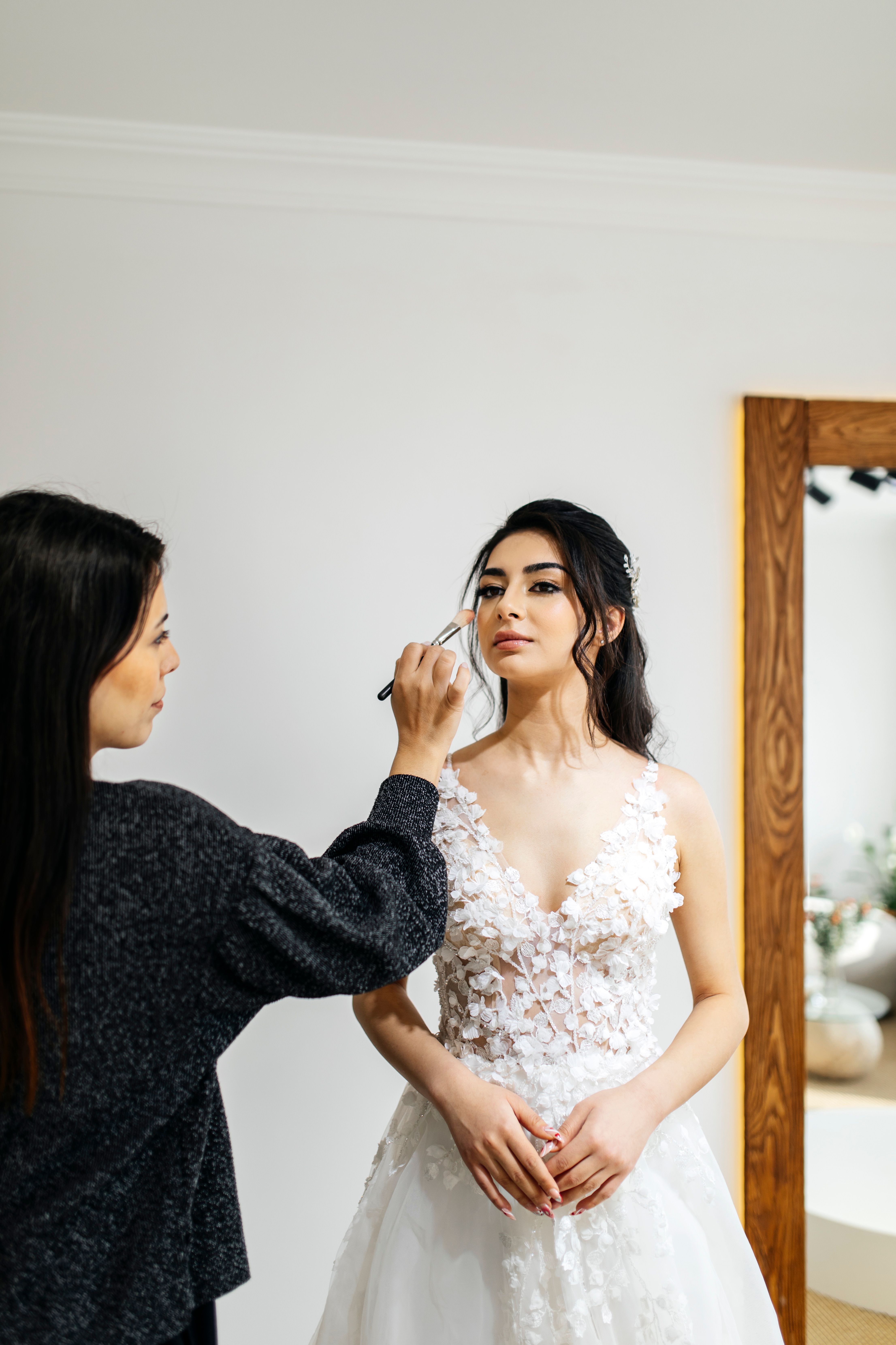 A bride in a beautiful gown being prepared by a beautician with elegant makeup indoors.