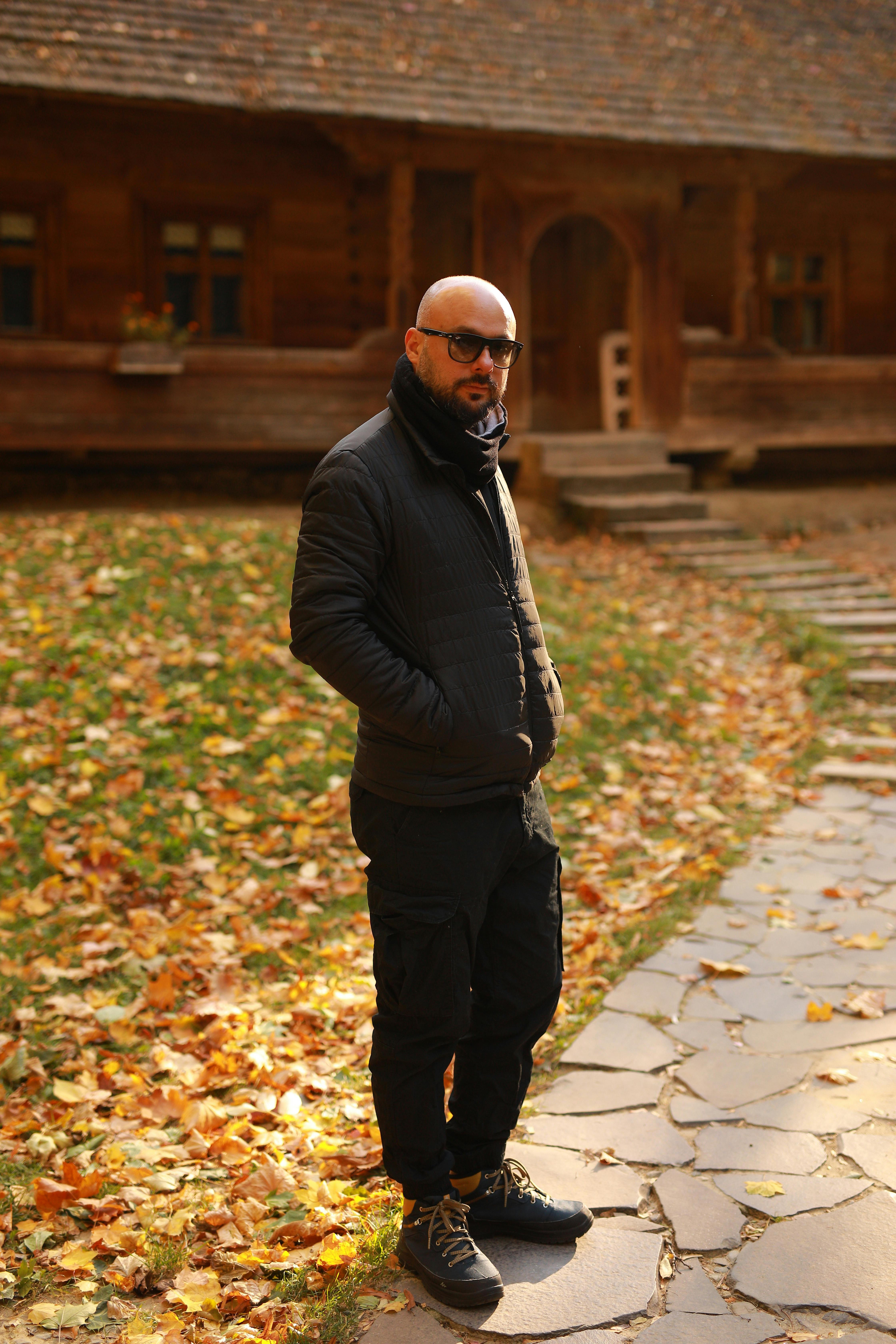 Man wearing autumn outfit standing on a leaf-covered path, posing in front of a rustic wooden house.
