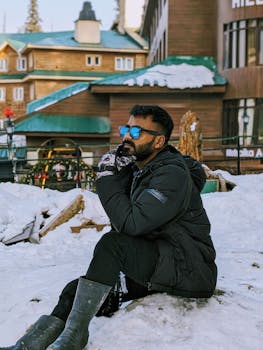 Man in winter attire sitting on snow in urban area, admiring the surroundings.