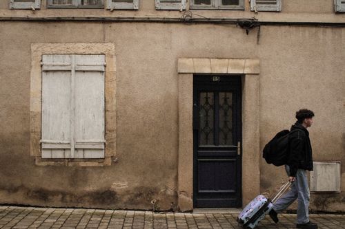 Free Man walking with luggage past a vintage building exterior with wooden shutters. Stock Photo