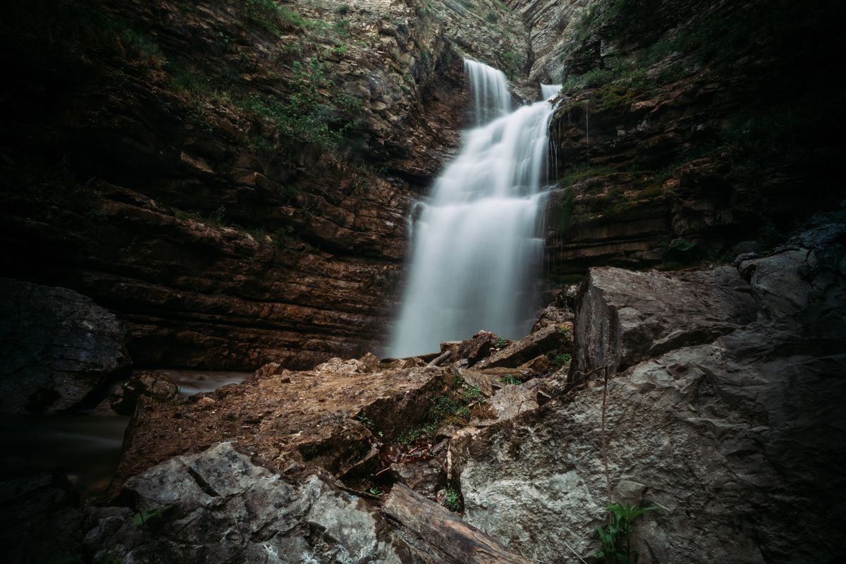 A waterfall flowing through a rocky gorge surrounded by green forest, viewed from a walking path